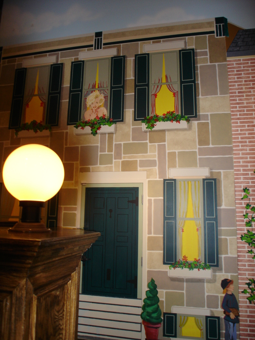 Corner brick building facade mural with green shutters and lamppost, car museum backdrop, Palm Beach, FL