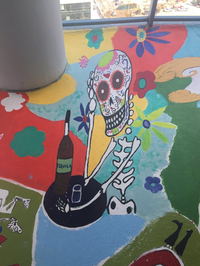 Wide view of the toasting skeleton scene beside a flower-crowned sugar skull face, exterior terrace mural, Palm Beach, FL