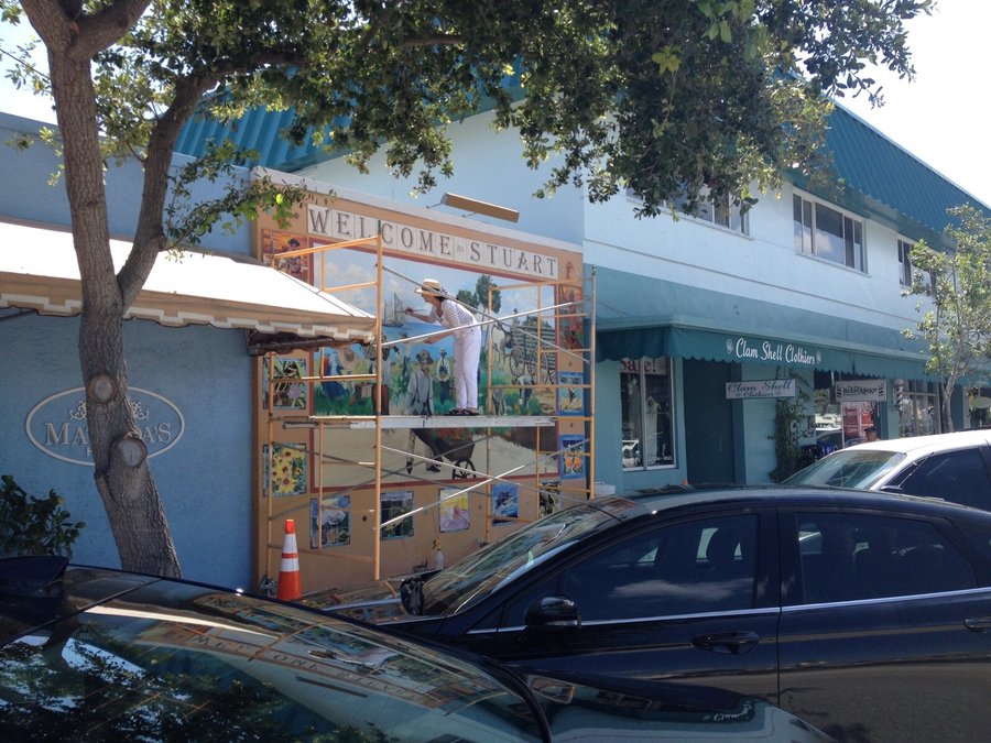 Distant street view of the Stuart mural on the building corner, storefronts and parked cars in foreground, Palm Beach, FL